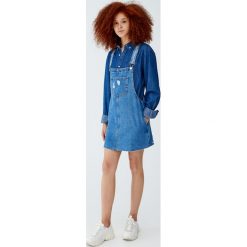 Jeansowa sukienka ogrodniczka z niewykończonym dołem. Niebieskie sukienki damskie z falbanami marki Pull&Bear, z jeansu. Za 89,90 zł.
