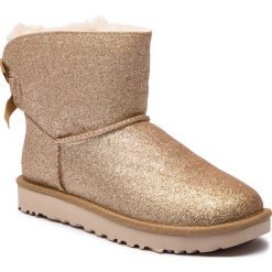 Buty UGG - W Mini Bailey Bow Sparkle 1100053 W/Gold. Żółte kozaki damskie marki Ugg, z tworzywa sztucznego, na niskim obcasie, bez zapięcia. W wyprzedaży za 569,00 zł.